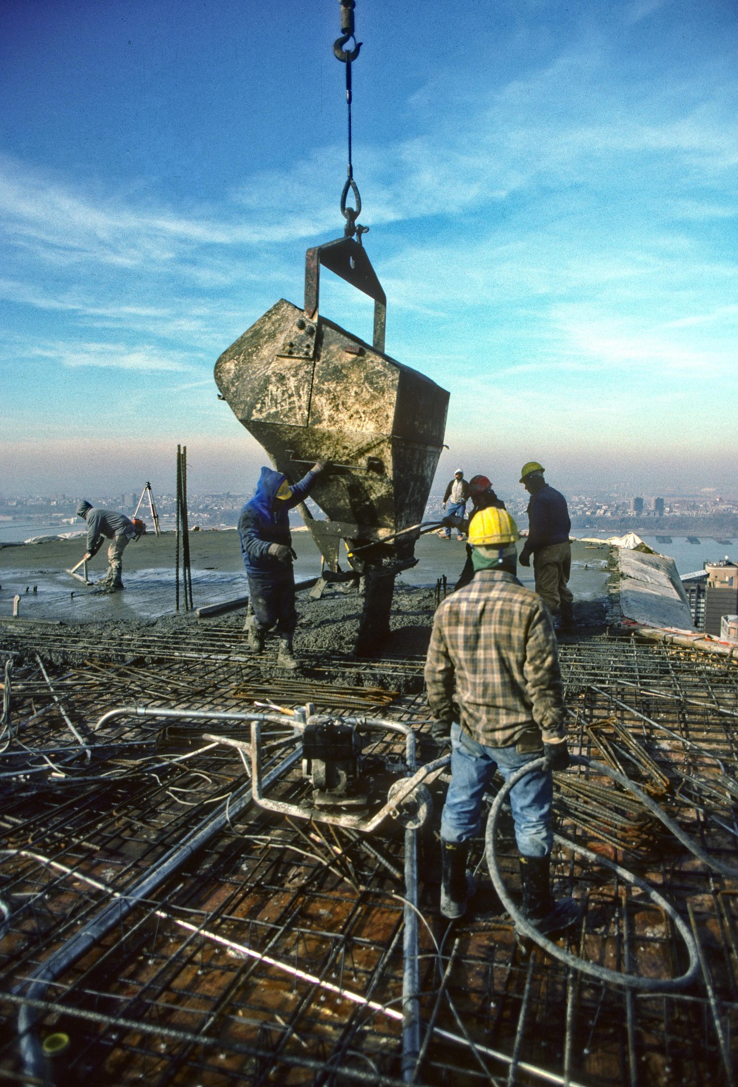 Crane lowering a concrete bucket for a pour, city skyline behind