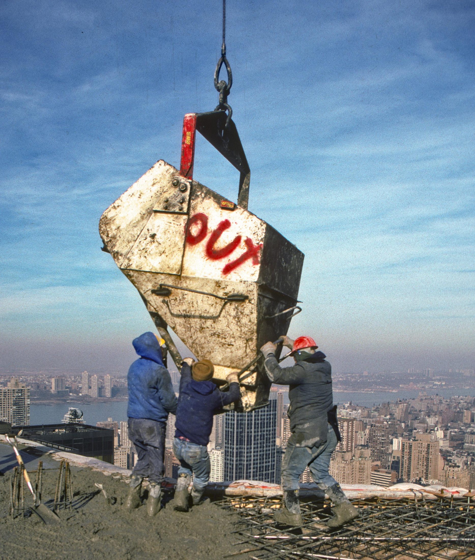 Workers guiding a concrete bucket marked OUT, city skyline behind