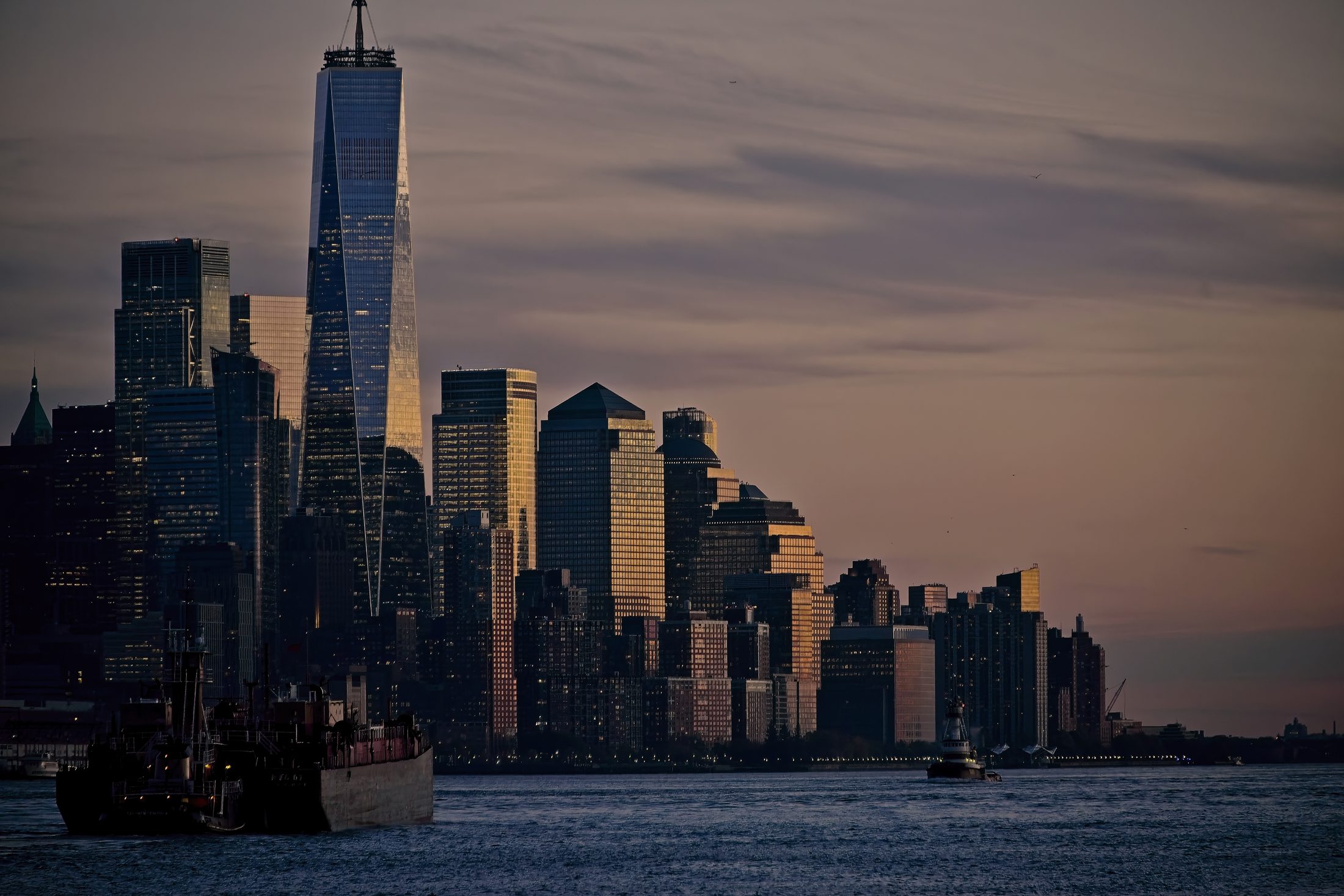 Lower Manhattan skyline at dusk from the harbor, tugboat and barge in the foreground